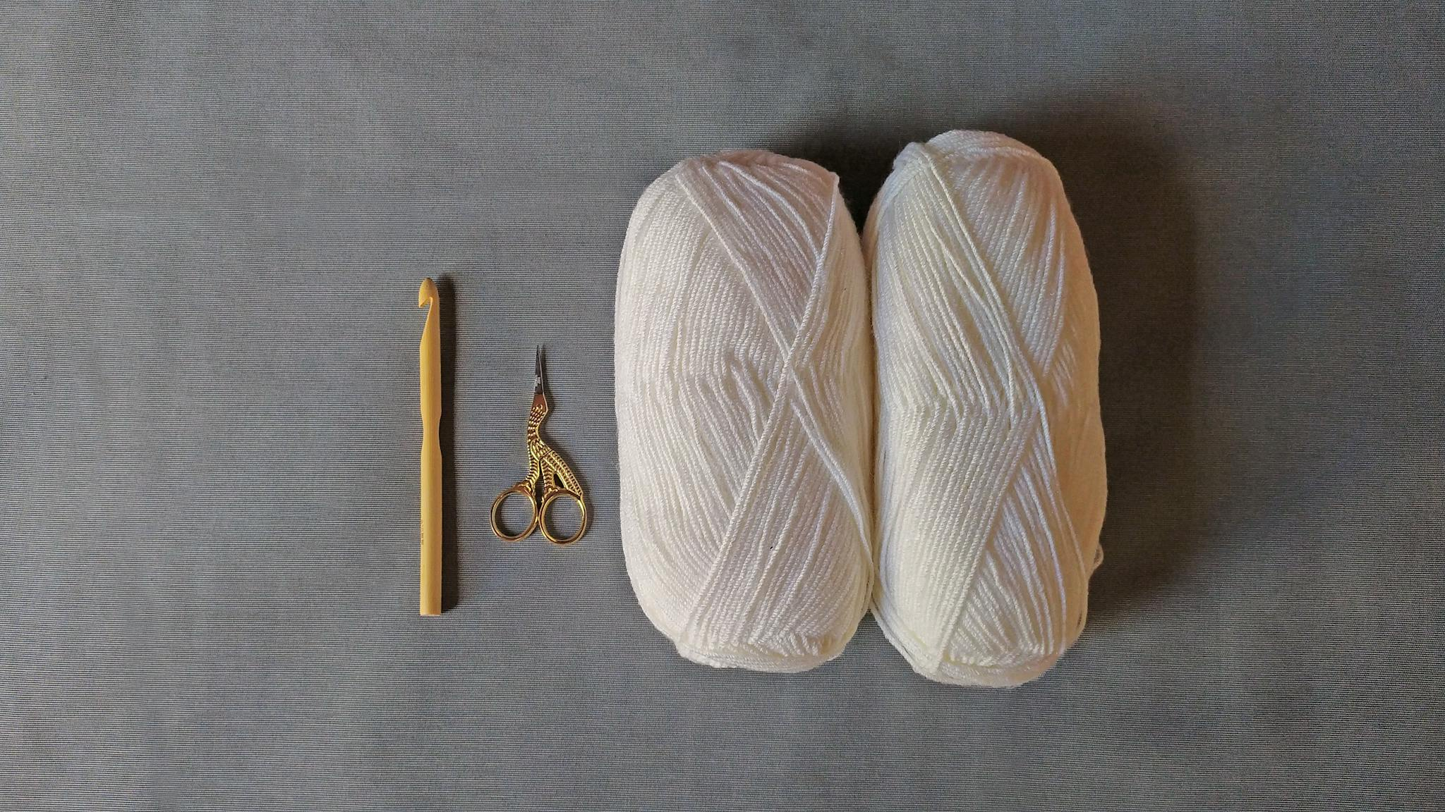 A minimalist flat lay featuring a crochet hook, scissors, and white yarn on a gray background.